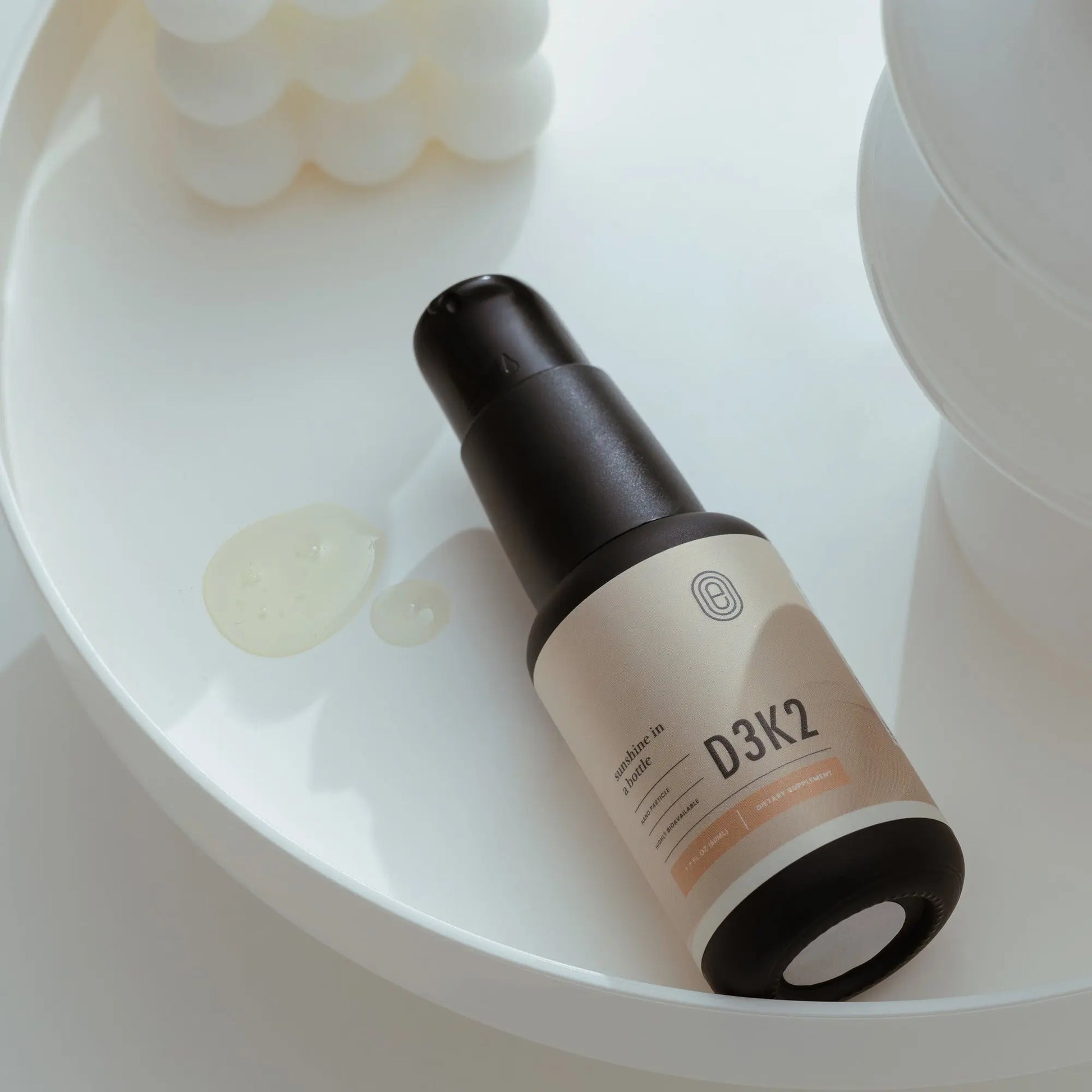 Emagene Life D3K2 supplement bottle on white tray with serum drops, minimalist setting